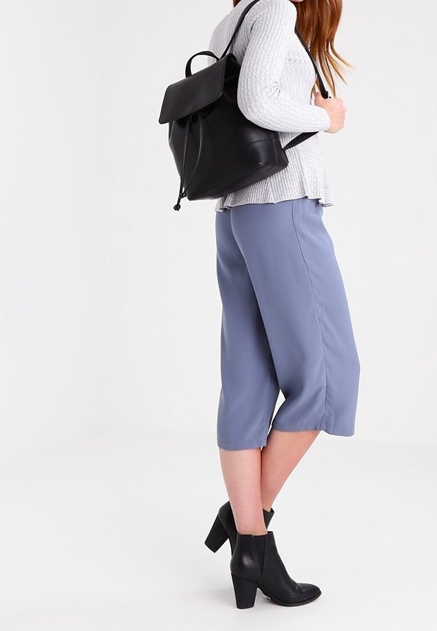 Femme portant un pull gris, un pantalon bleu court, des bottines à talons noires et un sac à dos en cuir noir sur le dos.