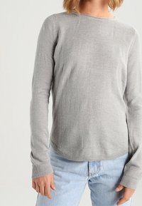 Pull gris à manches longues avec un col rond et des poignets côtelés, fabriqué en tissu doux et texturé, porté avec un jean en denim bleu clair.
