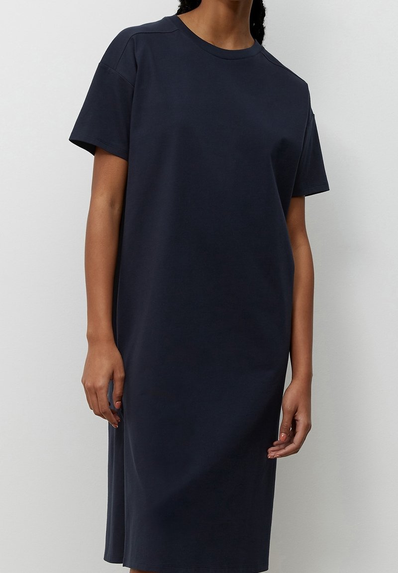 Femme portant une robe ample à manches courtes, bleu marine foncé, longueur genou, debout devant un fond blanc uni.