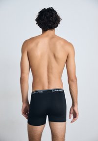 Schwarze Baumwollboxershorts mit einem Calvin Klein-Logo-Bund, getragen von einer Person, die einen nackten Rücken und Oberkörper vor einem hellen Hintergrund zeigt.