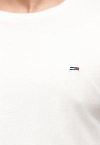Κοντινή λήψη ενός λευκού T-shirt με ένα μικρό ορθογώνιο λογότυπο σε ναυτικό μπλε, λευκό και κόκκινο στην περιοχή του πάνω μέρους του στήθους.