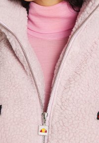 Różowa polarowa kurtka z suwakiem, o miękkim, teksturowanym wykończeniu. Posiada srebrny zamek błyskawiczny oraz haftowane akcenty na przodzie. Nosi się ją w warstwie z różowym golfem.