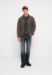 Brown zip-up jacket with a textured finish, black t-shirt underneath, blue faded jeans, and black shoes, topped with a red cap.