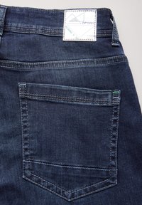 Jeans de mezclilla oscura con un bolsillo trasero que presenta un diseño de costura reforzada. Una etiqueta blanca en la cinturilla muestra patrones geométricos.