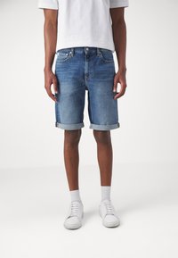 Denimshorts i medelblått med uppvikt fåll, med en klassisk femfickdesign och subtila blekningar. Bärs med vita sneakers.