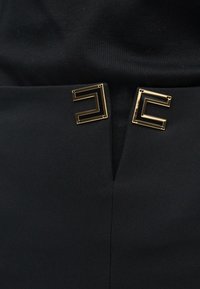 Κοντινό πλάνο μαύρου υφάσματος με δύο μεταλλικά γεωμετρικά κούμπωματα σε χρυσό και μαύρο, τοποθετημένα το ένα απέναντι στο άλλο κοντά σε μια κάθετη ραφή.