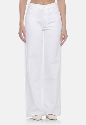 Donna che indossa pantaloni bianchi a vita alta e gamba larga e décolleté beige con punta a spillo, in piedi con le mani rilassate ai lati.