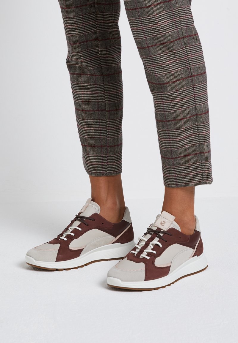 Zapatillas marrones y beige con materiales de ante y malla, que cuentan con cordones blancos, una suela de goma y un diseño aerodinámico.