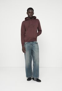 Kastanjefarvet hættetrøje med logo, iført sammen med brede, falmede blå denimjeans og sorte sko med hvide striber. Glat materiale, afslappet pasform.