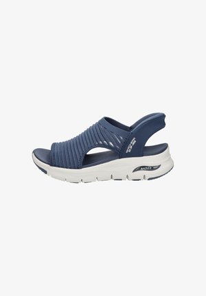 Navy blue slip-on sandal with stretchy knitted upper, cushioned white sole labeled "Arch Fit," and open heel strap for comfort and support.