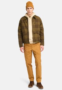 Brun og grønternet jakke over en beige hættetrøje, parret med orange bukser og brune støvler. Modellen har en sennepsfarvet beanie med et logo på.