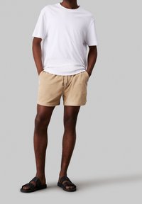 Beige froté šortky s elastickým pasem, doplněné bílým tričkem s kulatým výstřihem a černými sandály. Jednoduchý, pohodlný outfit.