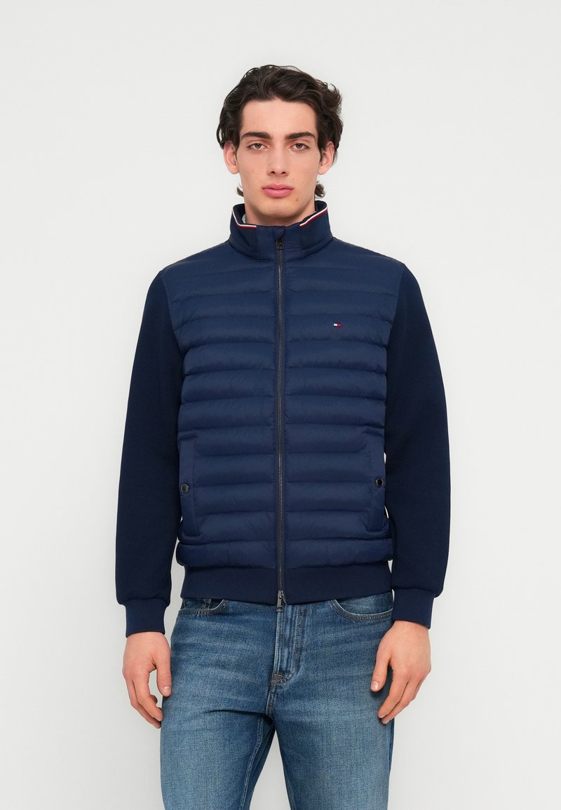Jeune homme portant une veste matelassée bleu marine avec fermeture éclair à l'avant et un jean, debout devant un fond blanc uni.