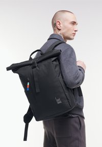 Czarny plecak typu roll-top z regulowanymi ramiączkami, kieszenią na zamek i detalem logo. Wykonany z trwałego materiału, charakteryzujący się minimalistycznym designem.