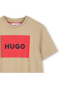 Μπεζ βαμβακερό T-shirt με ένα κόκκινο ορθογώνιο σχέδιο που φέρει τη λέξη "HUGO" με έντονα μαύρα γράμματα. Κοντά μανίκια και στρογγυλή λαιμόκοψη.