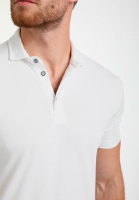 Camiseta polo blanca con un cierre de dos botones, mangas cortas y tejido texturizado. Detalle de logo sutil en el pecho.