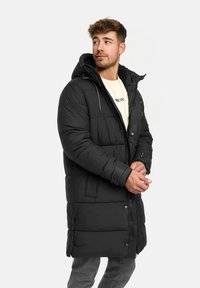 Zwarte gewatteerde lange jas met een capuchon, voorzien van een ritssluiting, twee zijzakken en een gestructureerde afwerking. Draagt over een lichtgekleurde blouse.