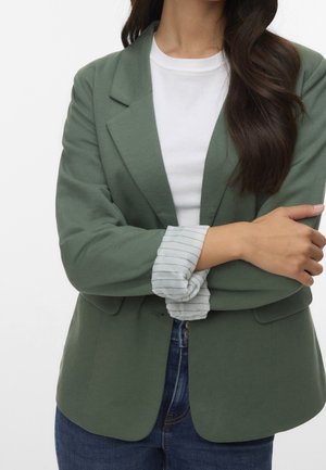 Femme aux longs cheveux foncés portant un blazer vert sur une chemise blanche et un jean bleu, les bras croisés avec les poignets de chemise rayés visibles.