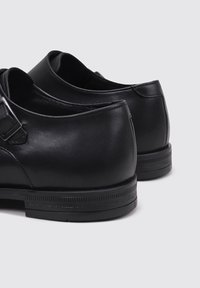 Zapatos de cuero negro con un acabado suave, punta redondeada y tacón bajo. Presentan un detalle de correa y hebilla simple. Suela de goma con agarre texturizado.