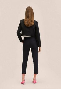 Zwarte cropped jas over een lichte top, gecombineerd met marineblauwe slim-fit broek en roze sandaaltjes met banden. Glad doek en getailleerde pasvorm zichtbaar.