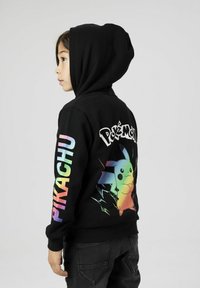 Felpa nera con un grafico colorato di Pikachu sul retro e la scritta "Pokémon". Maniche lunghe con "PIKACHU" in colori arcobaleno lungo un braccio.