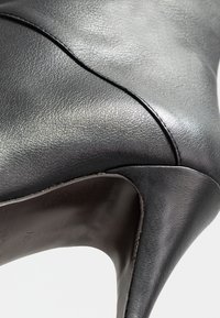 Černá vysoká kožená botička vyrobená z texturované kůže. Má špičatou špičku a zakřivený výřez, který ukazuje hladký, lesklý povrch.