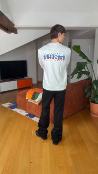 Mann steht drinnen mit dem Rücken zur Kamera, trägt ein weißes Sweatshirt mit dem Aufdruck "1985 Tommy Jeans", schwarze Hose, neben einem orangefarbenen Sofa und einer hohen grünen Pflanze.