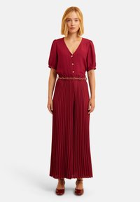 Burgunderfarbener plissierter Jumpsuit mit V-Ausschnitt und Puffärmeln, ausgestattet mit einer Knopfleiste und einem goldenen Kettenakzent um die Taille. Glatte Textur.