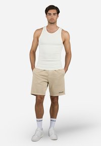 Hvit ribbestrikket tanktopp kombinert med beige shorts, med et logoaccent. Modellen har på seg hvite joggesko og hvite sokker med svart tekst.