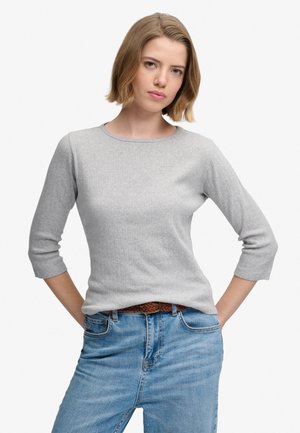 Kvinde med kort blondt hår iført en lysegrå sweater med trekvartærmer og blå jeans med et brunt flettet bælte.