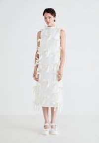 White sleeveless dress featuring textured fabric, adorned with numerous cream-colored bows, knee-length. Footwear includes white platform shoes.