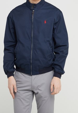 Homme portant une veste zippée bleu marine avec un logo rouge sur la poitrine et un pantalon gris clair, debout devant un fond uni.