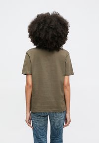 Asmuo su garbanotais plaukais stovi nugara, dėvi alyvuogių žalią trumpomis rankovėmis marškinėlį ir mėlynas džinsines kelnes su pakartojamu „JOOP! JEANS“ raštu.