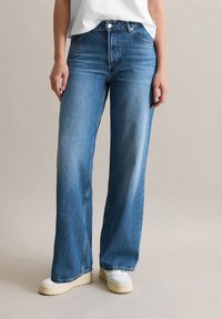 Helle blaue weite Jeans aus Denim mit hoher Taille, zwei Fronttaschen und Kontrastnähten. Kombiniert mit weißen Sneakern.