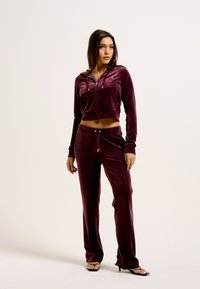 Tuta in velluto di un bordeaux scuro con felpa corta e pantaloni con coulisse. I dettagli notevoli includono cuciture a contrasto e accenti in hardware argentato.
