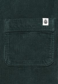 Tecido de corduroy verde escuro com um bolso frontal com textura de ribbing vertical e uma pequena etiqueta branca da marca no canto.