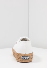 Sneaker Superga bianca in tela con una spessa suola in juta intrecciata, vista da dietro su una superficie bianca contro pareti bianche.