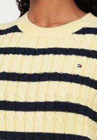 Pletený sveter s horizontálnymi pruhmi v žltej a námorníckej modrej farbe, textúrovaným šnúrovaním a malou nášivkou loga na hrudi.