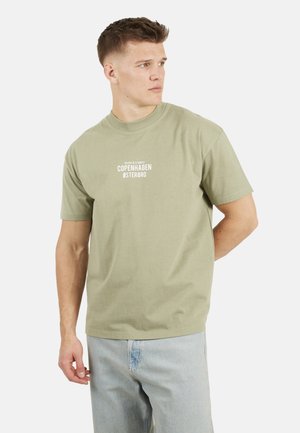 Jeune homme aux cheveux courts portant un T-shirt vert clair avec le texte "Copenhagen Østerbro" et un jean bleu clair, regardant vers sa droite.