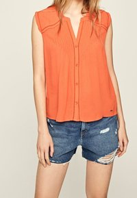 Ärmlös orange blus med plisserad detalj och knapplist fram. Matchad med slitna blå denimshorts med fransade sömmar.