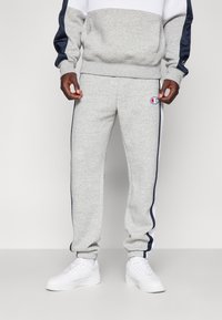Pantalones de chándal grises con rayas laterales en azul marino y un logo bordado. Hechos de tela suave, cuentan con puños acanalados y cintura con cordón.