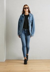 Giacca di jeans sopra una maglietta nera, abbinata a jeans skinny azzurri e sneakers nere. Modella con occhiali da sole, che cammina su un pavimento di legno.