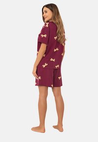 Kobieta stoi boso, ubrana w bordową bluzkę z krótkim rękawem i szorty w jasne żółte wzory w kokardki, zwrócona bokiem z głową przekręconą do tyłu.