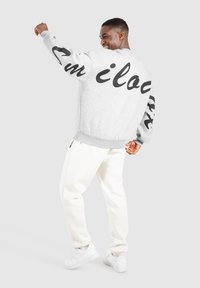 Sweatshirt gris avec texte noir sur les manches, présentant un col rond et des poignets côtelés. Associé à un pantalon de survêtement blanc et des baskets blanches.