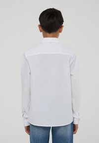 Camicia bianca a maniche lunghe con colletto, caratterizzata da una trama liscia e un orlo leggermente curvo, abbinata a jeans blu.