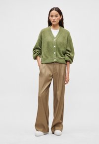 Πράσινη μάλλινη ζακέτα με V-λαιμόκοψη, φαρδιά μανίκια και μεγάλα κουμπιά, συνδυασμένη με μπεζ παντελόνια ψηλόμεσα και λευκά αθλητικά παπούτσια.