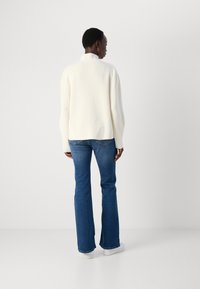 Cream ribbed sweater with a mock neck and relaxed fit, paired with blue flared jeans and white sneakers, viewed from the back.