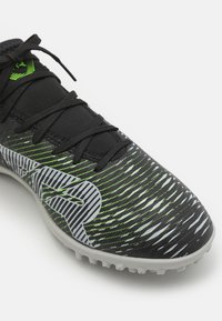 Zapatilla deportiva sintética negra con rayas diagonales verdes y blancas, puntera reforzada y cordones negros. Diseñada para ofrecer tracción en superficies de hierba.