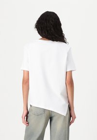 T-shirt bianco in cotone con maniche corte, scollatura rotonda e orlo posteriore asimmetrico. Abbinato a jeans in denim chiaro.