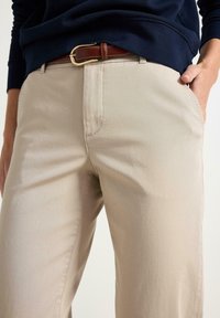 Beige bavlněné kalhoty s rovnými nohavicemi, ze předními kapsami a hnědým koženým pásem se zlatou sponou. Texturovaná látka s viditelným švem.